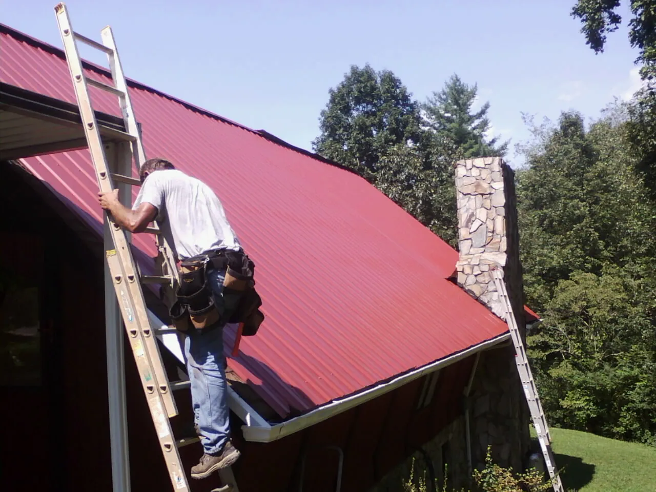 Roofer on a ladder beside a red metal roof and stone chimney — Soffit & Fascia Repair by Roofing Brothers in Bryant